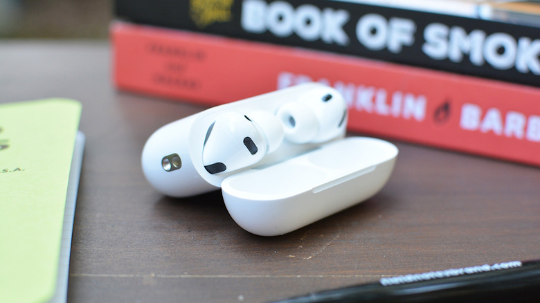 A pair of AirPods Pro 3 sit in their case on desk next to some books, a pen and notepad.